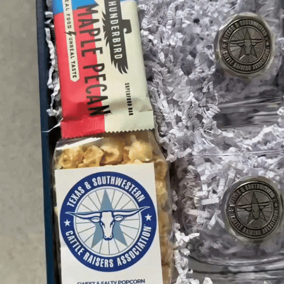 Close-up inside the delivered custom corporate gift box: medallion glasses, popcorn and a snack bar in white crinkle paper