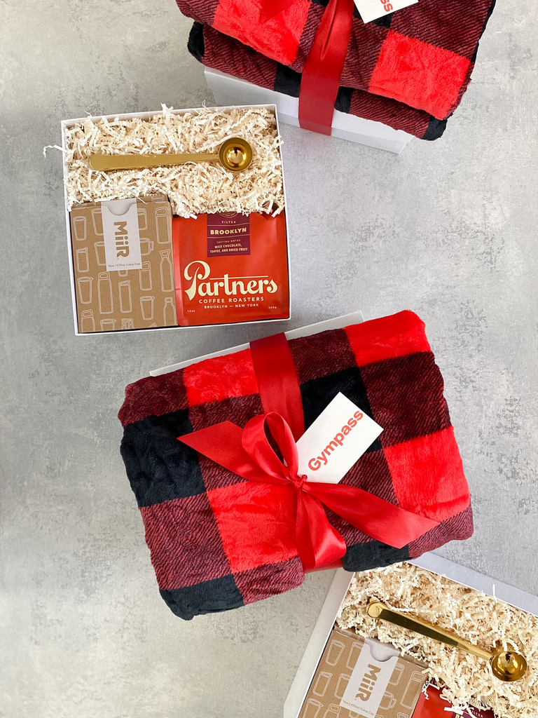 Corporate Christmas gift boxes with buffalo plaid throw blankets, coffee bags, and travel mugs tied in red ribbon
