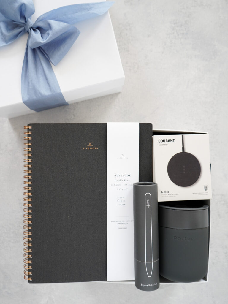 Open white gift box with blue ribbon containing an Appointed notebook, Squire rollerball pen, Courant wireless charger, and Porter travel mug