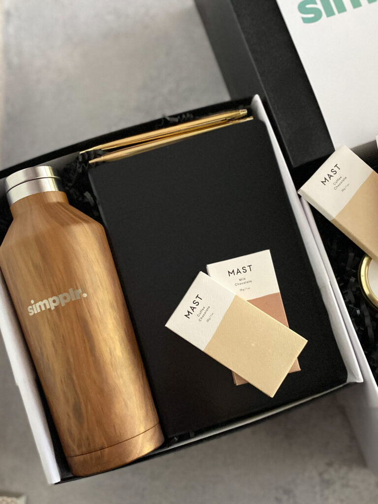Branded corporate gift set with a Simpplr logo water bottle, gold pen, black notebook, and Mast chocolate bars