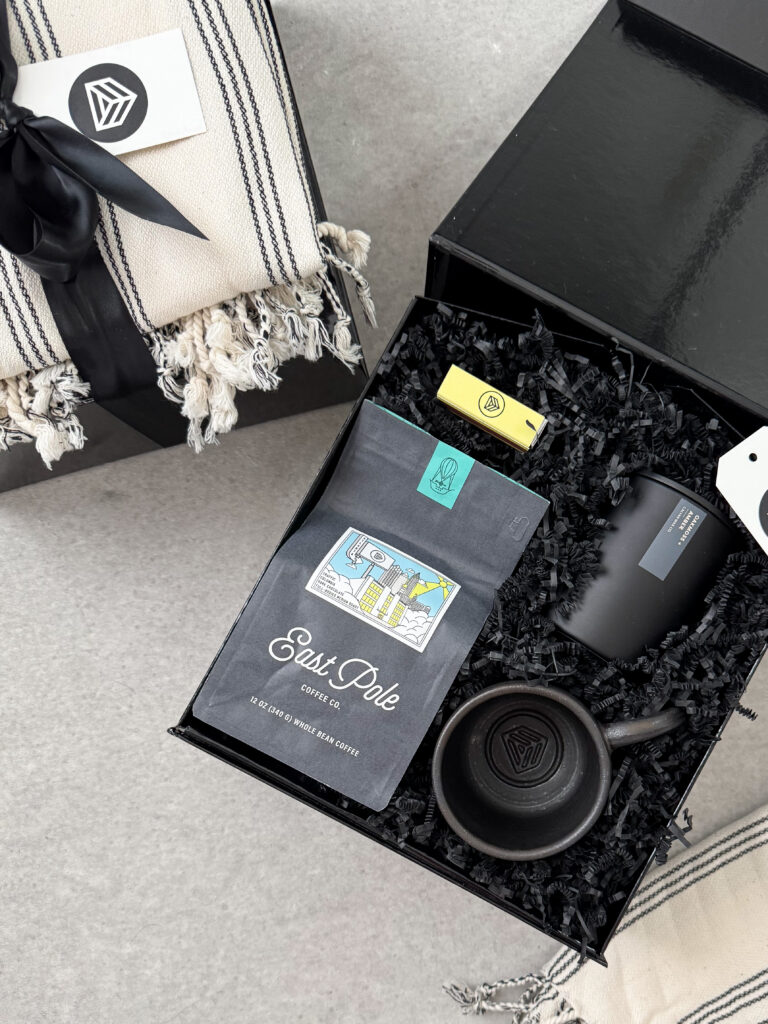 Black corporate Christmas gift box with coffee bag, mug, candle, and striped throw blanket wrapped in black ribbon