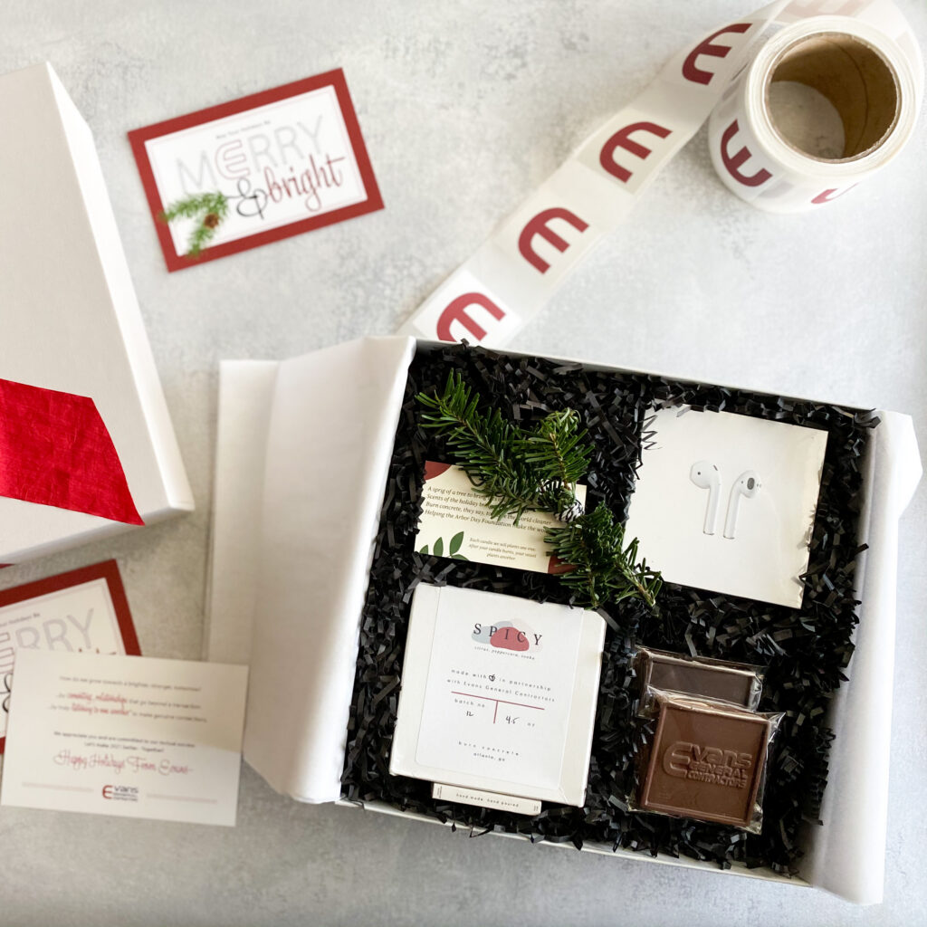 Alt text: Corporate Christmas gift box with wireless earbuds, spiced candle, branded chocolate, and pine sprig, "Merry & Bright" card alongside