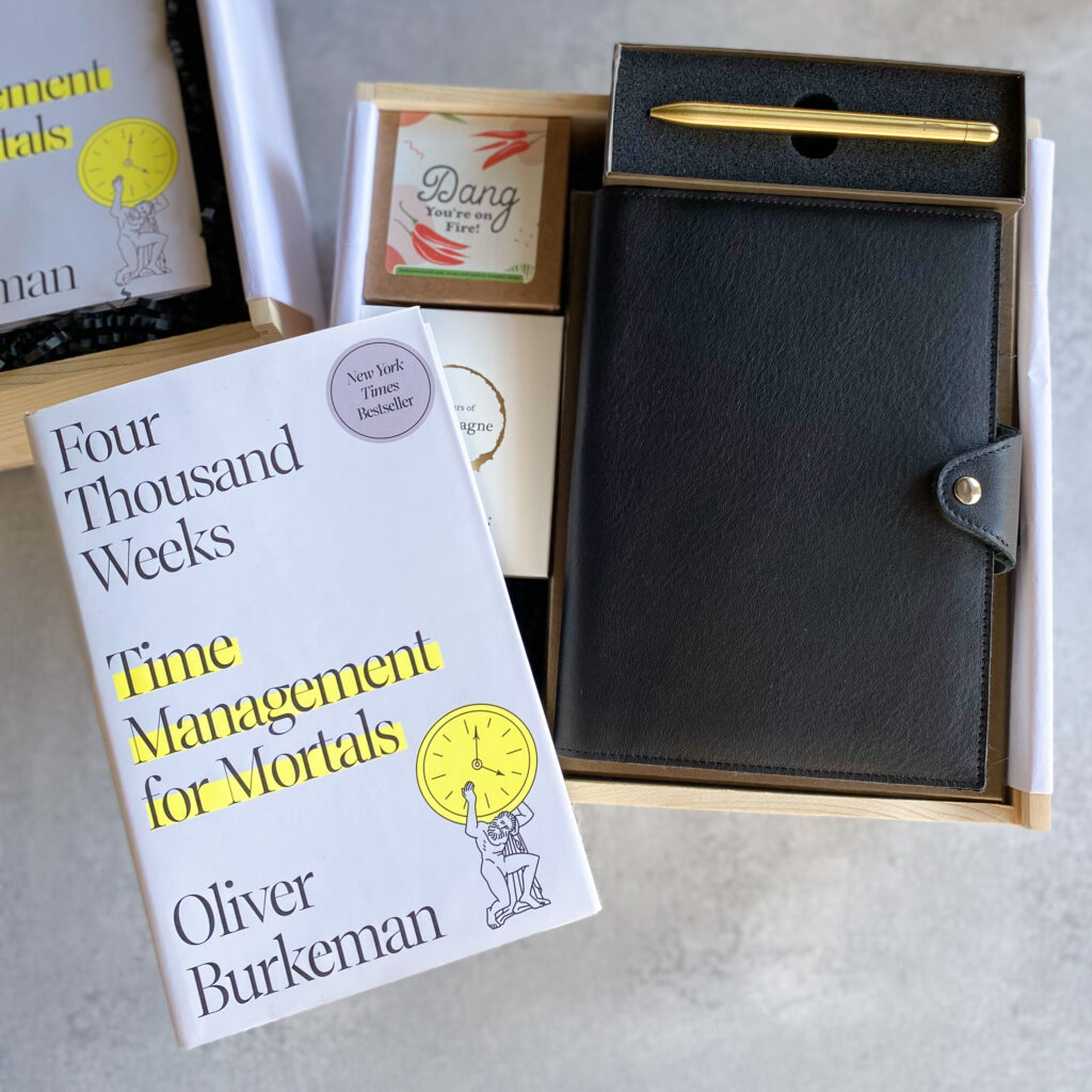 Corporate gift box with a black leather padfolio, gold pen, "Four Thousand Weeks" book by Oliver Burkeman, and small treat boxes
